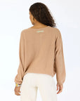A woman with wavy hair, facing away, wears a light brown Kerri Rosenthal Bassett Cardigan in featherweight cashmere with "LOVED" on the back neckline and light pants, standing against a plain white background.