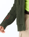 A person wears the Kerri Rosenthal Shawl Collar Helen Cardigan in green over a neon green shirt and khaki pants. The cardigan's sleeve features red cursive embroidery, with their relaxed hand visible.