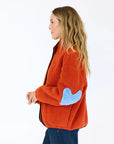 A woman with long wavy hair wears the cozy Kerri Rosenthal Coze Magoze cotton fleece jacket in orange with a blue heart elbow patch, standing sideways against a plain white background.