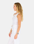 A woman with long blonde hair stands in profile against a white background, wearing the KR Tank by Kerri Rosenthal and white pants, accessorized with bracelets and a watch, showcasing effortless style.