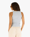 A person with long, wavy hair wears sunglasses and the Kerri Rosenthal KR Tank—a black cotton top with an embroidered heart—paired with beige pants. Shown from the back, hands in pockets, against a plain white background.