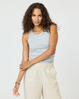 A woman with long curly hair wears the Kerri Rosenthal KR Tank in gray with light beige pants, a gold necklace, and a watch. She stands against a plain light background, looking at the camera and touching her arm.