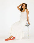 A woman in a white sleeveless top and the Kerri Rosenthal Vacance Skirt with lace trim sits on a painted stool, touching her hair and smiling. She wears orange slip-on shoes and sunglasses against a plain white background.