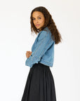 A woman with long curly hair models the Kerri Rosenthal Denim Jacket featuring heart elbow patches, paired with a black skirt. She stands sideways, looking at the camera against a plain white background.