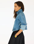 A woman with wavy hair wears a Kerri Rosenthal Denim Jacket, featuring heart elbow patches, paired with a black skirt and yellow watch, standing in profile against a plain white background.