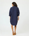 A woman with curly hair faces away, wearing the Kerri Rosenthal Gigi Denim Dress—a loose-fitting, navy blue, knee-length cotton shirt dress with three-quarter sleeves—and tan high-heeled sandals against a plain white background.