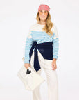 Wearing the Kerri Rosenthal Artist Long Sleeve Stripe Tee in blue and white, a woman pairs it with white pants, a navy sweater at her waist, glasses, and a large white bag with a black heart, all set against a plain white background.