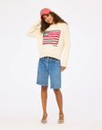 A woman smiles against a white background in the Kerri Rosenthal Roll Neck / US of Love Sweater, denim knee-length shorts, strappy sandals, and a red baseball cap—a perfect patriotic look.