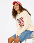 A smiling woman with long curly hair wears the Kerri Rosenthal Roll Neck / US of Love Sweater, denim shorts, and a coral pink cap as she sits on a paint-splattered stool against a plain white background.