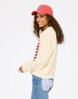 A smiling woman with curly hair wears a pink cap, blue jeans, and the Roll Neck /US of Love Sweater by Kerri Rosenthal. She stands sideways, looking at the camera against a plain white background.