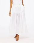 A woman in a sleeveless white top and a Kerri Rosenthal Vacance Pintuck Skirt with sheer details and tiered lace trim, paired with tan heeled sandals, stands against a plain white background.