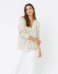 A woman with long, dark hair, wearing a Kerri Rosenthal Slouchy Cardigan in light beige made of 100% cotton, white pants, and featuring a pink heart appliqué on her left side, stands against a plain white background. Her right arm is bent, and her left arm is folded across her body.