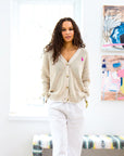 A person with long curly hair stands in a bright, white-walled room wearing the Slouchy Cardigan by Kerri Rosenthal and white pants. Colorful abstract art pieces are mounted on the wall behind them, with daylight streaming in through a window.