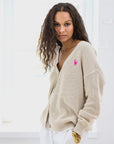 A person with long wavy hair is wearing a Kerri Rosenthal Slouchy Cardigan with a pink heart appliqué on the chest, and white trousers. They are standing indoors in a brightly lit room, looking towards the camera with a neutral expression.