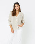 A woman with long brown hair wears a beige, 100% cotton Slouchy Cardigan from Kerri Rosenthal, adorned with a heart appliqué over a white shirt and white pants. She stands against a plain white background, her left hand in her pocket and wearing a relaxed expression.
