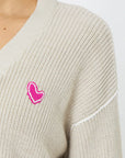Close-up of a person wearing the Kerri Rosenthal Slouchy Cardigan in light beige, featuring a pink heart-shaped appliqué on the left side. The focus is on the upper left part of this 100% cotton cardigan, highlighting its texture and the embroidered heart design.