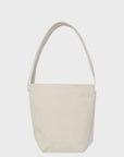 The Uma by Kule is a minimalist beige canvas bucket bag made from 100% cotton, featuring a single shoulder strap. It's showcased against a plain white background with a simple design and no visible pockets or embellishments.