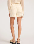 A person faces away wearing BROCHU WALKER's KAIA EMBROIDERED SHORT in cream Italian fabric, paired with a beige knit sweater and platform sandals, against a plain light background.