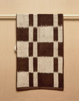 The Karin Hand Towel by Autumn Sonata, made from organic cotton and featuring a bold brown and beige plaid pattern inspired by Ikat weaving, hangs neatly on a rod against a beige background.
