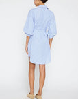 A woman with her hair tied up models the BROCHU WALKER KATE BELTED DRESS, a light blue shirtdress with blouson sleeves and white pinstripes, paired with beige slip-on shoes against a plain white background.
