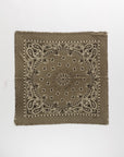 The Hachiko Scarf by KUJTEN is a square olive green bandana in printed cashmere, showcasing white and black paisley and floral designs, frayed edges, and a textured finish. It’s presented on a plain white background.