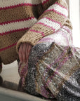 A person sits California cool wearing the Le Superbe Nirvana Sweater—a chunky knit with brown, white, and pink stripes plus a jeweled neckline—one hand on their knee beside a sequined skirt.