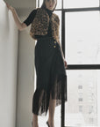 A woman with straight brown hair stands by a large window, confidently wearing the Le Superbe Utility Fringe Skirt, styled with a leopard print vest, black top, and black heels.