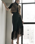 A woman poses indoors by a large window, wearing the Le Superbe Treasure Vest in leopard print over a black top, paired with a fringe-detailed black skirt and black high-heeled shoes.