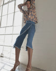 A person poses indoors near large windows, exuding stylish confidence while wearing the Le Superbe Its a Wrap Pant paired with a long-sleeve floral blouse and heeled ankle-strap sandals.