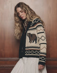 The Bear Lodge Cardigan by The Great Inc. styled with a white skirt, modeled by a wood wall for a cozy look.