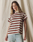 A person with wavy, light brown hair wears The Boxy Crew F25 tee by The Great Inc. with red, green, and white stripes and light blue jeans, standing before a neutral textured fabric backdrop.