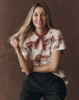 A woman with long blonde hair smiles against a brown background, wearing The Boxy Crew 100% cotton t-shirt by The Great Inc., featuring horses, plus a red neck scarf and dark pants, while holding a strand of her hair.