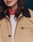 A person wears The Great Inc. Barn Jacket, a tan garment-dyed cotton workwear piece with a dark brown collar, over a white shirt and red patterned bandana. Only the lower half of their face and upper torso are visible.