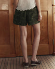 Someone stands indoors on a wooden floor, wearing The Dolphin Short by The Great Inc.—green camo cotton sateen shorts—with a white sleeveless top and brown boat shoes. Their upper body is mostly out of the frame.