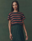 A woman with long braided hair wears The Boxy Crew F25 by The Great Inc., a 1970s-inspired vintage striped tee, paired with a high-waisted dark green lace skirt, standing against a muted green background.
