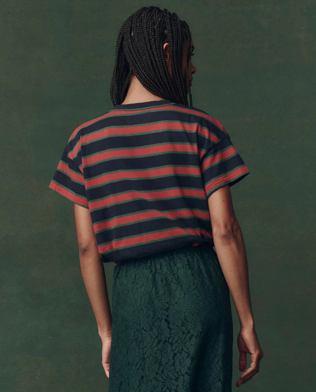 A person with braided hair faces away, wearing The Great Inc.'s The Boxy Crew F25 vintage striped tee in dark hues and a textured dark green skirt, set against a muted green background.