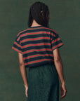 A person with braided hair faces away, wearing The Great Inc.'s The Boxy Crew F25 vintage striped tee in dark hues and a textured dark green skirt, set against a muted green background.