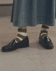 Black loafers and Collegiate Sock by The Great Inc. paired with a long denim skirt on concrete flooring.
