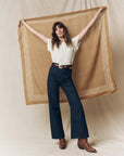 A woman smiles with arms outstretched, holding The Big Bandana by The Great Inc. behind her while wearing a white T-shirt, dark blue wide-leg jeans, and brown boots against a neutral backdrop.