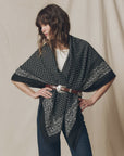 A woman stands with her hands on her hips against a neutral fabric backdrop, wearing The Big Bandana by The Great Inc. as an oversized shawl over a white top, cinched at the waist with a brown leather belt.