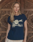 A woman with long blonde hair wears The Great Inc.'s navy blue The Boxy Crew w/ Everglade Graphic tee and a cream skirt, standing by an ornate wooden door and looking at the camera—an effortlessly chic Los Angeles style.