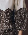 A close-up of The Chariot Top, showcasing its textured cotton with black lace patterns over white fabric and elaborate, symmetrical designs on the sleeve and bodice.