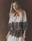 A woman with long blonde hair wears The Chariot Top—a white western lace print top—with a red scarf around her neck, standing against a brown background.