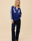 A blonde woman wears the DayDreamer Los Angeles Stars Rugby, an oversized blue long sleeve shirt with "L A" on the front, paired with dark jeans. She stands against a plain beige background, looking forward with a slight smile.
