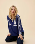 A blonde woman kneels on the floor against a plain beige background wearing the DayDreamer Los Angeles Stars Rugby long sleeve shirt with "LA" and star-striped sleeves.