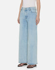 A person wears FRAME's Le Slim Palazzo light blue wide-leg jeans made from a sustainable cotton blend paired with a white top, standing casually. These high-rise jeans with pockets are stylishly completed with black sandals.