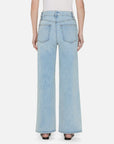 A person stands against a white background in FRAME's Le Slim Palazzo jeans, featuring a high-rise fit, light blue hue, sustainable cotton blend, wide-leg palazzo silhouette, and two back pockets. They pair the jeans with black heels for an effortlessly chic look.