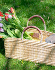 The Faire Gardener Long Basket, handmade in Morocco from ethically-sourced woven palm leaves, rests on green grass with red and purple tulips and a wooden-handled gardening tool. Soft sunlight casts gentle shadows across the scene.