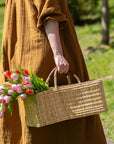 A person in a brown dress stands outdoors on a sunny day, holding the Faire Gardener Long Basket—handmade in Morocco—by its rectangular handle, filled with pink and red tulips.
