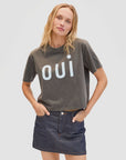 A woman with long blonde hair wears the Clare Vivier Le Carre Tee, a dark gray 100% cotton boxy shirt with "oui" in large white letters, paired with a dark denim skirt. She stands against a plain light background, hand in her pocket.